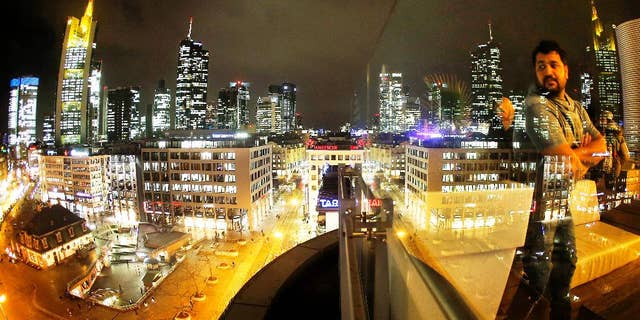 The buildings of the banking district are reflected in a glass railing while a man watches from a skyline outdoor restaurant in Frankfurt, Germany, Wednesday, Jan. 4, 2017. (AP Photo/Michael Probst)