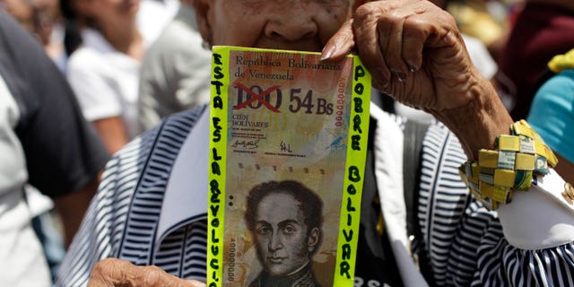 FILE - In this Feb. 23, 2013 file photo, an anti-government protester in Caracas holds up a fake Venezuelan banknote that reads in Spanish, "This is the revolution. Poor Bolivar," with the value of 100 crossed out and 54 written next to it. Venezuelans have been scrambling for dollars as their bolivar currency is in free fall, selling on the black market for a tenth its official value. In response, the government is trying to block access to websites that Venezuelans use to track the black market exchange rate. (AP Photo/Ariana Cubillos, File)