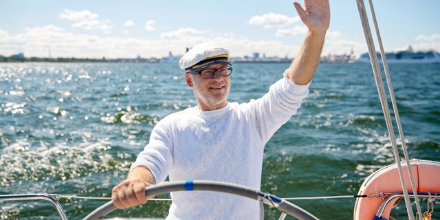 sailing, age, tourism, travel and people concept - happy senior man in captain hat on steering wheel and waving hand sail boat or yacht floating in sea