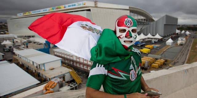 Backdropped by the Itaquerao stadium, a Mexico soccer fan poses for a photo wearing a wrestling mask and a representation of his country's national flag as a cape, in Sao Paulo, Brazil, Tuesday, June 10, 2014. The high-profile inaugural match will be played at the troubled Itaquerao, which was one of the most delayed venues for the World Cup. It's already known that the stadium's roof won't be fully finished until after the tournament. (AP Photo/Eduardo Verdugo)