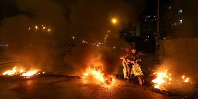 Lebanese citizens ride their scooter between burning tires that were set on fire by Sunni protesters, at one of the major roadways between Beirut and the capital's southern suburbs, a Hezbollah stronghold, to show their support with Sunni town of Arsal, in Beirut, Lebanon, Tuesday, March 18, 2014. Gunmen from Lebanon's militant Hezbollah group and local Shiite Muslim residents tightened their blockade of a Sunni town of Arsal near the Syrian border Tuesday, sparking concerns that thousands of Syrian refugees stranded in the area could be cut off from humanitarian aid. (AP Photo/Hussein Malla)