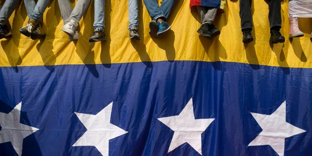 People sit on a wall draped by a Venezuelan flag during a protest against President Nicolas Maduro in Caracas, Venezuela, Wednesday, Oct. 26, 2016. Venezuela's standoff deepened after congress voted to open a political trial against Maduro for breaking the constitutional order and opposition leaders called for mass demonstrations on Wednesday to drive the leader from office. (AP Photo/Alejandro Cegarra)