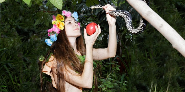 Young girl with floral wreath on her head holding an apple in one hand and a snake in the other. Foliage forest on dark background. Temptation of Eve.