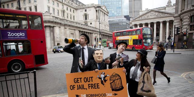 Campaigners from the Robin Hood Tax organization dressed as bankers, including one wearing a mask representing British Chancellor of the Exchequer George Osborne, hold placards as they pose for the media outside Mansion House, backdropped by the Bank of England at left, in London, Wednesday, June 10, 2015. The group say their idea for a Robin Hood Tax is a tiny tax on the financial sector that could generate billions of pounds annually to fight poverty and climate change. The stunt was held by the group to coincide with Osborne's annual economic speech due to be held at Mansion House on Wednesday evening.  (AP Photo/Matt Dunham)