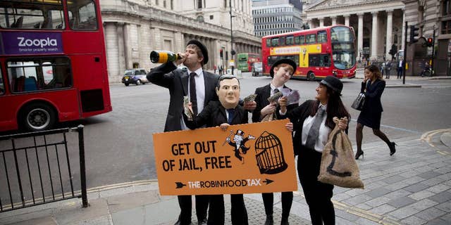 Campaigners from the Robin Hood Tax organization dressed as bankers, including one wearing a mask representing British Chancellor of the Exchequer George Osborne, hold placards as they pose for the media outside Mansion House, backdropped by the Bank of England at left, in London, Wednesday, June 10, 2015. The group say their idea for a Robin Hood Tax is a tiny tax on the financial sector that could generate billions of pounds annually to fight poverty and climate change. The stunt was held by the group to coincide with Osborne's annual economic speech due to be held at Mansion House on Wednesday evening. (AP Photo/Matt Dunham)