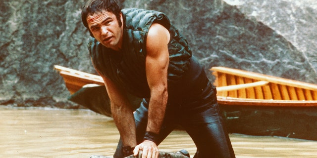 Burt Reynolds, US actor, holding onto some rocks as he struggles in the water with his boat behind him in a publicity still issued for the film, 'Deliverance', 美国. 的 1972 惊悚片, directed by John Boorman, starred Reynolds as Lewis Medlock.