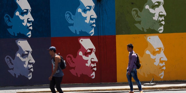Two men skate board past a mural of Venezuela's late President Hugo Chavez in downtown Caracas, Venezuela, Tuesday, March 4, 2014. Hes been dead a year, but Chavezs face and voice are everywhere. On Wednesday, the country will mark Chavez's death, who died at age 58 on March 5, 2013. (AP Photo/Rodrigo Abd)