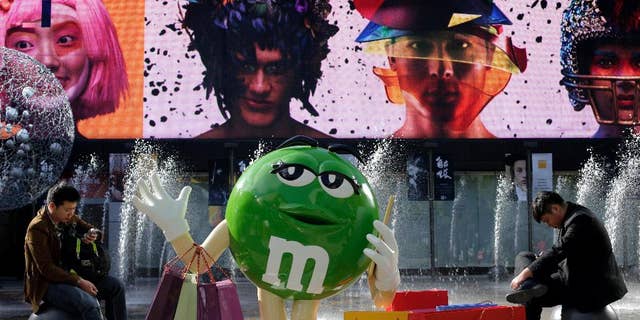 Chinese men browse their mobile phones near a M&M chocolates figure on display at a shopping mall in Beijing, Wednesday, Oct. 28, 2015. Facing pressure to pep up a cooling economy, Chinese leaders met on Monday and will announce a five-year plan effort to shift China from reliance on trade and investment to more self-sustaining growth driven by domestic consumption. The Chinese words on the bag reads "Don't worry how much you shop, just spend to release stress" (AP Photo/Andy Wong)