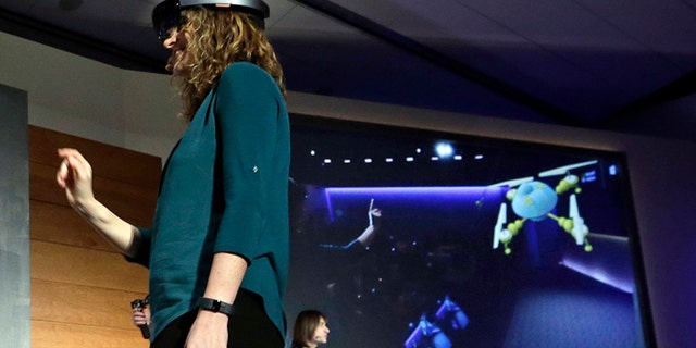 In this photo taken Wednesday, Jan. 21, 2015, Microsoft's Lorraine Bardeen demonstrates a hologram device as what she "sees" is projected on a screen behind during an event at the company's headquarters in Redmond, Wash. With the new HoloLens headset, Microsoft is offering real-world examples to show how you might use three-dimensional digital images _ or holograms _ in daily life. (AP Photo/Elaine Thompson)