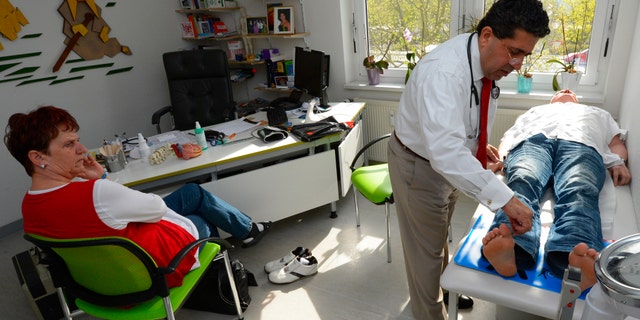 SCHWEDT, GERMANY - APRIL 30: Medical doctor Amin Ballouz checks the reflexes of the lower legs of a patient as the patient's wife looks on during a medical examinaiton at Ballouz's practice on April 30, 2013 in Schwedt, Germany. Ballouz was born in Lebanon and moved to Germany as a child, and has had a general practitioner's practice in the small, east German town of Schwedt since 2010. Many of his patients are elderly and live in small villages in the region around Schwedt and Ballouz travels daily in one of his five Trabant cars to pay housecalls. Eastern Germany faces a chronic shortage of country doctors to serve rural communities. (Photo by Theo Heimann/Getty Images)