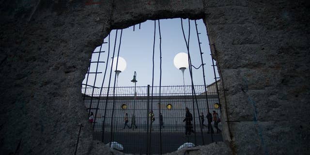 People pass by balloons of the art project 'Lichtgrenze 2014' (lit. 'lightborder 2014') photographed through a hole in remains of the berlin Wall in Berlin, Germany, Friday, Nov. 7, 2014. The light installation featuring 8,000 luminous white balloons commemorates the division of Berlin where the 25th anniversary of the fall of the wall is marked with numerous events on the weekend. (AP Photo/Steffi Loos)
