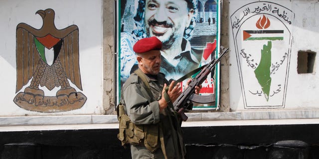 A member of the Palestine Liberation Organization's Armed Struggle unit, that is in charge of security in the Palestinian camps, holds his AK-47 machine gun as he walks in front of a portrait of the late Palestinian leader, Yasser Arafat, at a checkpoint in the Palestinian refugee camp of Ein el-Hilweh, in Sidon, Lebanon, Tuesday June 19, 2012. Palestinians refugees are protesting after clashes broke out Monday in the Nahr el-Bared camp in the north of Lebanon during the funeral of a Palestinian who was killed in a confrontation Friday. The Arabic logo painted on the wall, right, reads "The Palestine Liberation Organization, national unity, liberation." (AP Photo/Mohammed Zaatari)