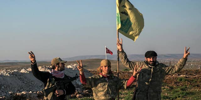In this Sunday, Feb. 22, 2015, Syrian Kurdish militia members of YPG make a V-sign next to a poster of Abdullah Ocalan, jailed Kurdish rebel leader, and a Turkish army tank in the background in Esme village in Aleppo province, Syria.