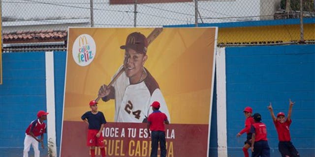 Two boys stand in front of an advertisement featuring Miguel Cabrera during a baseball practice in Maracay.