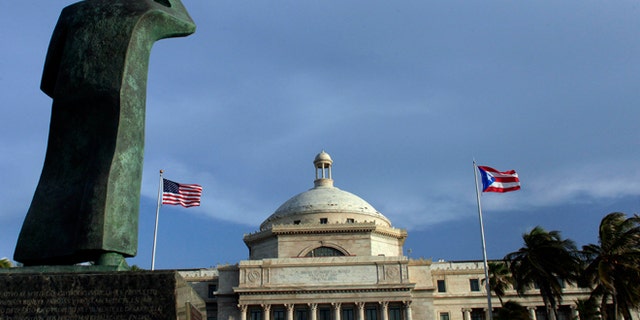 FILE - In this Wednesday, July 29, 2015, file photo, a bronze statue of San Juan Bautista stands in front of Puerto Ricoâs Capitol as U.S. and Puerto Rican flags fly in San Juan, Puerto Rico. After months of pleading from the government of Puerto Rico, the U.S. Congress agreed on Wednesday, May 18, 2016, to help the territory restructure its massive public debt. (AP Photo/Ricardo Arduengo, File)