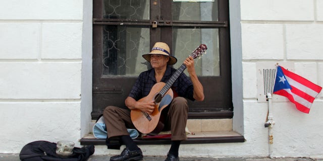 A man plays his guitar while he begs for money in front of a closed down business in Old San Juan, Puerto Rico, Monday, June 29, 2015. International economists released a critical report on Puerto Rico's economy Monday on the heels of the governor's warning that the island can't pay its $72 billion public debt. (AP Photo/Ricardo Arduengo)