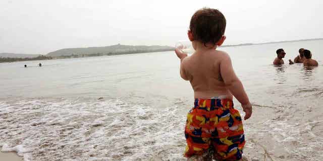 In this May 23, 2015 photo, 17-month-old Hunt Van Benschoten drinks from a bottle as he stands in the water at a beach in San Juan, Puerto Rico. No matter how experienced a traveler you are, an overseas trip taken with a baby or toddler requires changes in planning, expectations and itineraries. (Amber Hunt via AP)