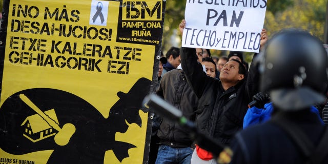 Nov. 14, 2012: A man who identified himself as Edwin, 32, from Ecuador, right, who was evicted from his home over debts, holds up banner reading ''We need bread, roof and employment'' beside a poster to support people fighting against evictions reading, ''No More Evictions'', in front of Spanish riot police during a general strike against government austerity measures, in Pamplona, northern Spain.