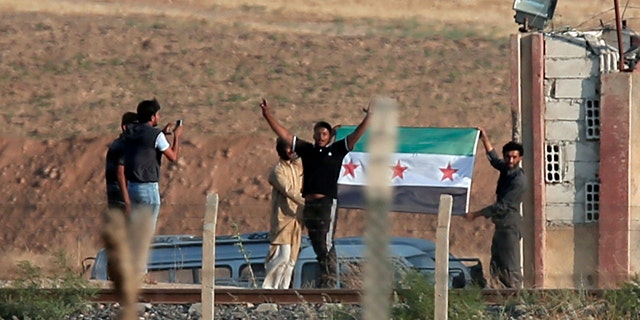 June 15, 2015: In this photo taken from the Turkish side of the border between Turkey and Syria, in Akcakale, southeastern Turkey, Kurdish people wave a Free Syria Army group flag in the outskirts of Tal Abyad, Syria. (AP Photo/Lefteris Pitarakis)