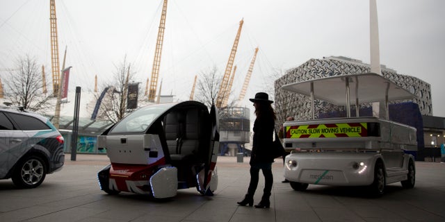 A woman poses for photographers beside a prototype driverless car called a LUTZ Pathfinder Pod, center, and a Meridian shuttle, right, during a launch event for the media near the O2 Arena in London.