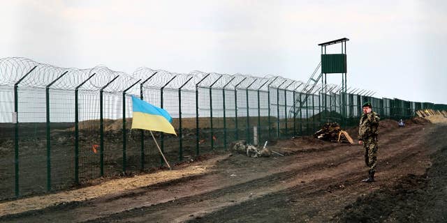 In this photo taken on Saturday, April 18, 2015, a Ukrainian border guard officer speaks on a phone near a national flag attached to the fence on the Ukrainian-Russian border near Hoptivka, Kharkiv region, eastern Ukraine. The flimsy, razor wire-topped fences popping up along bare expanses of Ukraine’s eastern frontier are the first line of defense against a much-feared Russian invasion. (AP Photo/Inna Varenytsia)