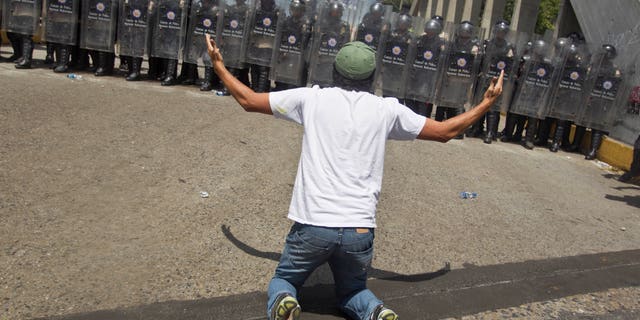 An anti-government demonsrator kneels in front of a formation of Bolivarian National Police in riot gear, during clashes at the Central University of Venezuela, UCV, in Caracas, Venezuela, Thursday, March 20, 2014. Thursday dawned with two more opposition politicians, San Cristobal Mayor Daniel Ceballos and San Diego Mayor Enzo Scarano, behind bars. Police used tear gas and water cannons to disperse a student-called protest of several thousand people in Caracas, some of those demonstrating against the arrests of the mayors. (AP Photo/Esteban Felix)