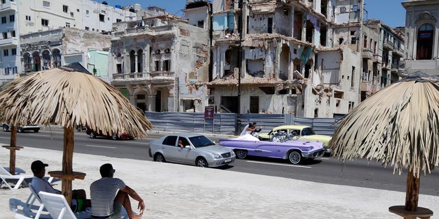 People sit on deck chairs on an artificial beach as a bride and groom ride a classic American convertible car on the Malecon in Havana, Cuba, Thursday, May 21, 2015. The artificial beach is part of a collective artistic intervention named Â¨Behind The WallÂ¨for the Havana Biennial Art Fair which starts on May 22. (AP Photo/Desmond Boylan)