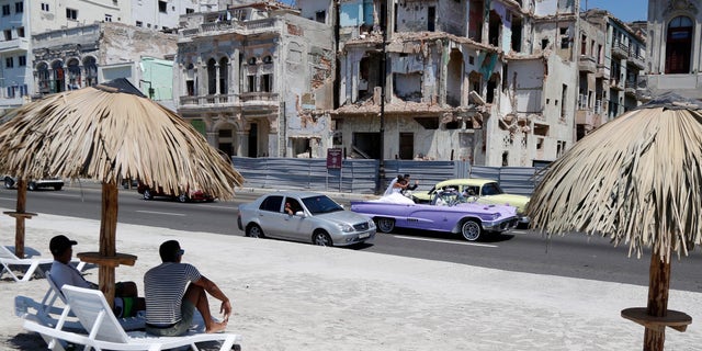 People sit on deck chairs on an artificial beach as a bride and groom ride a classic American convertible car on the Malecon in Havana, Cuba, Thursday, May 21, 2015. The artificial beach is part of a collective artistic intervention named ¨Behind The Wall¨for the Havana Biennial Art Fair which starts on May 22. (AP Photo/Desmond Boylan)