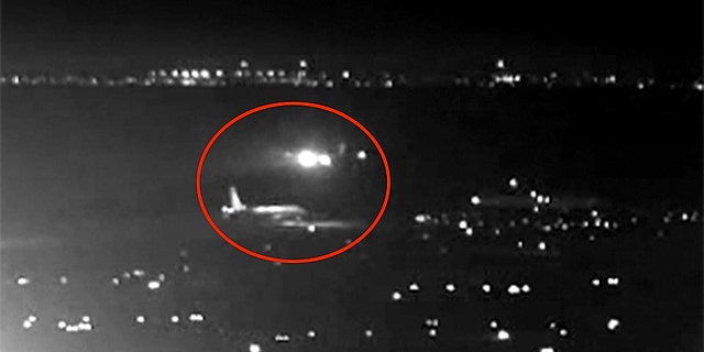 An Air Canada plane can be seen flying just above a United Airlines flight waiting on the taxiway.