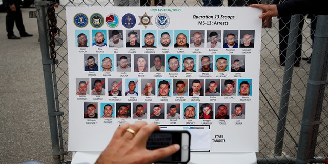 A reporter uses his smartphone to photograph a board showing images of MS-13 gang members during a news conference Wednesday, May 17, 2017, in Los Angeles. Hundreds of federal and local law enforcement fanned out across Los Angeles, serving arrest and search warrants as part of a three-year investigation into the violent and brutal street gang MS-13. (AP Photo/Jae C. Hong)