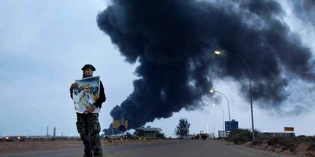 March 12, 2011: A pro-Gadhafi fighter holds a poster of Libyan Leader Moammar Gadhafi as black plume of smoke rises from the burning oil refinery as they are pictured during a government-organized visit for foreign media in Ras Lanouf, Libya