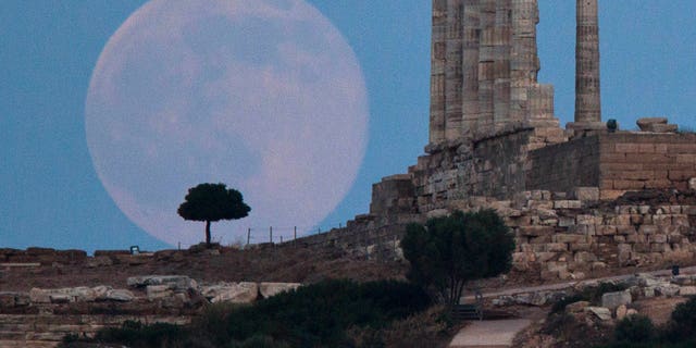 The full moon rises behind a tree next to the ruins of the ancient Temple of Poseidon near Athens, Greece. 