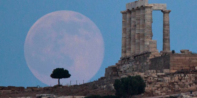 The full moon rises behind a tree next to the ruins of the ancient Temple of Poseidon near Athens, Greece.