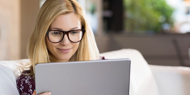 Happy young blonde woman sitting on sofa and using digital tablet. Happy young student with eyeglasses watching a movie on tablet. Female student studying on tablet at home.