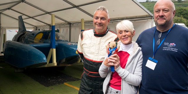 Hydroplane Pilot Ted Walsh, left, Gina Campbell the daughter of pilot Donald Campbell and engineer Bill Smith pose for a photo with the restored Bluebird K7 before it takes to the water for the first time in more than 50 years off the Isle of Bute on the west coast of Scotland, Saturday Aug. 4, 2018. The famed jet boat Bluebird has returned to the water for the first time since a 1967 crash that killed pilot Donald Campbell during a world speed-record attempt. Watched by Campbell's daughter Gina Campbell, the restored Bluebird was lowered Saturday into Loch Fad on Scotland's Isle of Bute, where it will undergo low-speed tests.(David Cheskin/PA via AP)