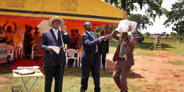 In this photo taken on Saturday, April 20 2013 and released by the Uganda Presidency, Uganda's President Yoweri Museveni, left, and State Minister for Youth and Children Affairs Ronald Kibuule, center, hand over what the president's office said was a sack of money containing 250 million Ugandan shillings - about $100,000 - to an unidentified member of a partisan group of youths, right, in Kaliro district east of the capital Kampala, in Uganda. It’s become an infamous photo in Uganda. The country’s leader looks on in bemusement as a young man bends low to carry a sack filled with a donation from the president _ almost $100,000 in cash. The money is a donation from President Yoweri Museveni to a partisan group of youths in eastern Uganda, where he struggles to win votes in national elections, and activists and opposition politicians are seizing upon the moment as a blatant example of political corruption in Uganda. The picture, which was distributed by the president’s office after the event last weekend, was meant to highlight Museveni’s generosity toward a group of youth. Instead it has focused attention on Museveni’s profligacy during political tours and what some say is his role in fueling graft in the East African country that he has led since 1986. (AP Photo/Uganda Presidency)