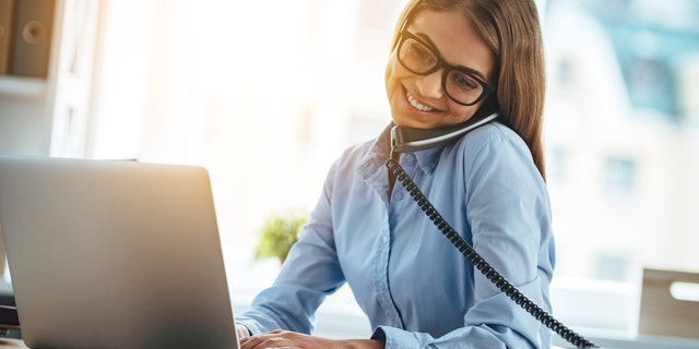 Cheerful young beautiful woman in glasses talking on the phone and using laptop with smile while sitting at her working place