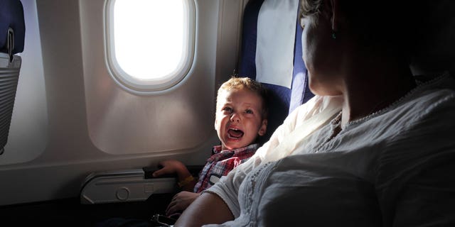 A child cries on an airplane while sitting in the seat next to his mother.