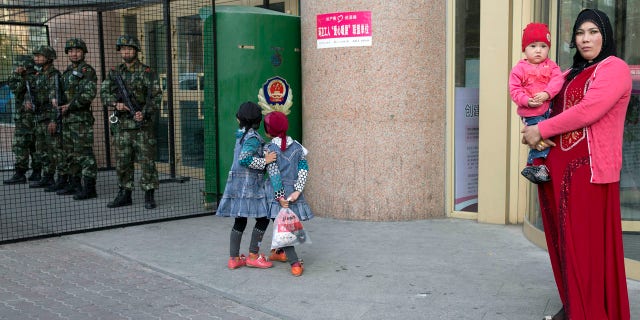 In this Thursday, May 1, 2014 file photo, an Uighur woman carries a toddler as children play near a cage protecting heavily armed Chinese paramilitary policemen on duty in Urumqi in northwest China's Xinjiang Uygur Autonomous Region.