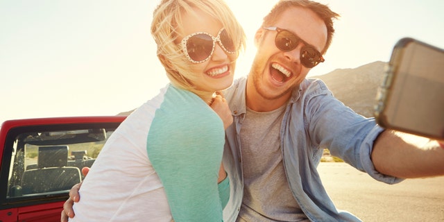 Couple On Road Trip Sit On Convertible Car Taking Selfie