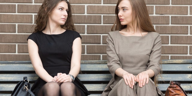 Portrait of two beautiful young female rivals sitting side by side on bench and looking at each other with challenging expressions. Attractive caucasian office women ready for confrontation