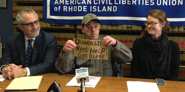 Michael Monteiro, center, with his lawyer Marc Gursky, left, and outreach worker Megan Smith, right, holds the sign that he used to solicit donations from a median strip in Cranston, R.I., during a news conference Wednesday, Dec. 16, 2015 in Providence, R.I. Monteiro said he has periodically asked for donations at the intersection for years, but stopped this summer after he was cited by police, then told by a judge not to return. Monteiro is now suing the city for violating his constitutional rights. (AP Photo/Michelle R. Smith)