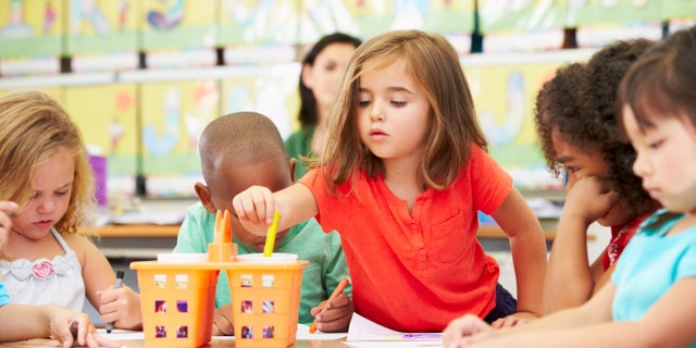 Group Of Elementary Age Children In Art Class With Teacher Reaching For Crayon