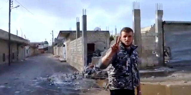 In this image taken from video obtained from the Ugarit News, which has been authenticated based on its contents and other AP reporting, A Free Syrian Army soldier flashes the victory sign, as damages caused by barrels fired from warplanes and rocket launchers is seen at background in Hama, Syria, on Monday, Jan. 28, 2013. (AP Photo/Ugarit News via AP video)
