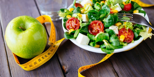 Fitness salad and apple fruit surrounded by measuring tape on rustic wooden table. Mixed greens, tomatos, diet cheese, olive oil and spices for healthy lifestyle concept.