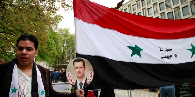 A Syrian supporting the regime of Syrian President Bashar Assad, holds a placard with his picture and the national flag as he participates in a protest outside the US Embassy in central London, Saturday, May 18, 2013. (AP Photo/Lefteris Pitarakis)