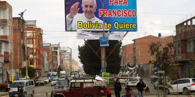 FILE - In this April 16, 2015, file photo, street food vendors set up their stand under a billboard featuring Pope Francis with a message that reads in Spanish; "Pope Francis, Bolivia loves you," in El Alto, Bolvia. Pope Francis' three-nation trip to South America that includes Ecuador, Bolivia and Paraguay, is set for July 6-12, though he will only spend six hours in Bolivia's capital due to the altitude, church officials say. (AP Photo/Juan Karita, File)