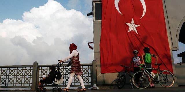 A woman passes with her child by two cyclist as they stand by a giant Turkey flag at the Galata bridge in Istanbul, on Thursday, Aug. 4, 2016. The Turkish government characterizes the movement of Fethullah Gulen, who lives in self-imposed exile in Pennsylvania, as a terrorist organization. (AP Photo/Petros Karadjias)
