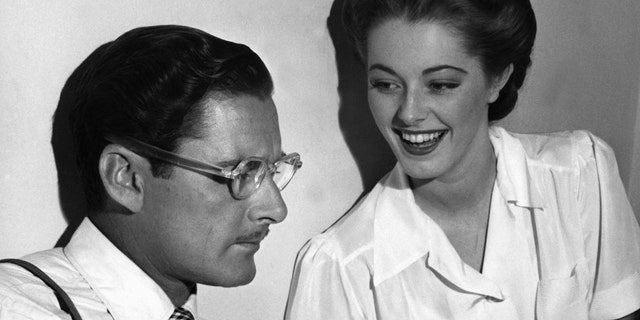 Errol Flynn seems to be in a literary mood as, between takes on the set of "Never Say Goodbye," he reads an amusing passage from a new novel to his leading lady, Eleanor Parker, who appears to enjoy the presentation in Los Angeles.