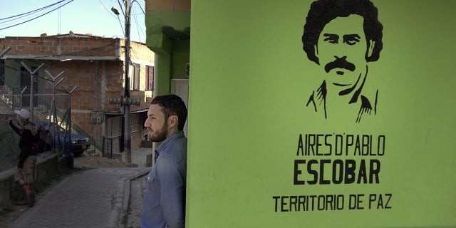 Doug Laux leans against a wall in the barrio with a stencil of Pablo Escobar?s face spray-painted on its surface.